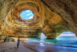 Praia Benagil Cave no Algarve com luz solar entrando pelo teto circular, iluminando a areia dourada e águas turquesa cristalinas.
