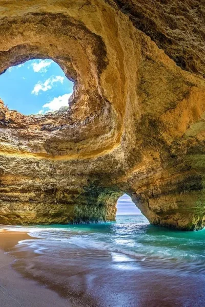Praia Benagil Cave no Algarve com luz solar entrando pelo teto circular, iluminando a areia dourada e águas turquesa cristalinas.
